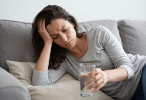 Early signs of dehydration - Woman feeling dizzy on sofa reaching for water showing when dehydration becomes more serious.