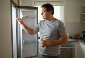 Mid-aged man opening a fridge holding a beer, illustrating lifestyle factors behind a beer gut
