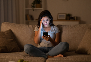 Woman sitting on a couch late at night using her phone, illustrating habits that can interfere with weight management