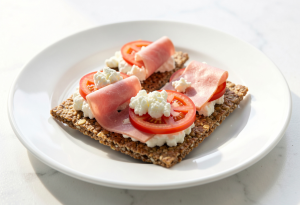 Two whole-grain crispbreads topped with cottage cheese, tomato slices, and lean ham on a plate, ready-to-eat snack