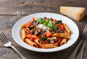 Plate of whole-grain spaghetti or penne with Bolognese sauce, vegetables, and Parmesan, garnished with fresh basil. Extra sauce and vegetables on the side for topping.