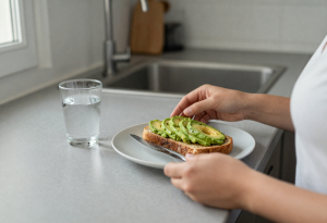 Person eating a small bowl of nuts and avocado toast at a kitchen counter, representing practical consumption of healthy fats in daily life.