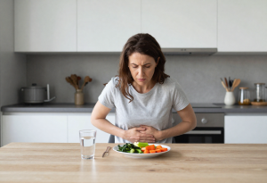 Woman at a kitchen table holding her stomach, with reflux-friendly foods and water nearby, illustrating causes of acid reflux