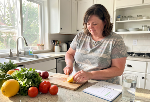 Woman at a sunlit kitchen counter cutting lemon for water, with prepped vegetables and a habit-tracking notebook nearby, illustrating daily routines to manage weight during menopause