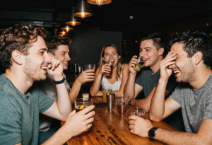 Group of young adults socializing at a bar, with one person lightly rubbing their eyes showing subtle fatigue or redness, illustrating short-term effects of alcohol on eye appearance
